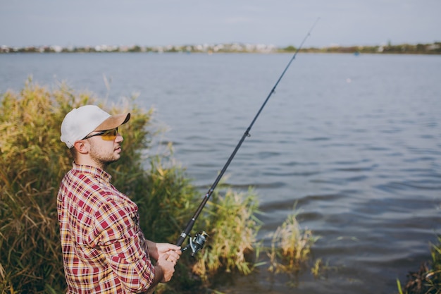 Side view Young unshaven man with a fishing rod in checkered shirt, cap and sunglasses looks into distance on lake from shore near shrubs and reeds. Lifestyle, recreation, fisherman leisure concept.