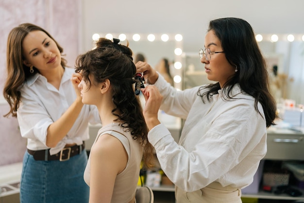 Side view of young woman being given applying highlighter for facial sculpting from female makeup artist Hairdresser curling hair using hair iron Concept of makeup and styling in 4 hands