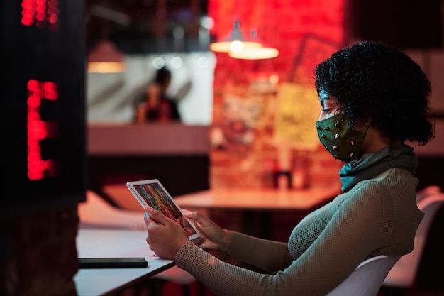 Photo side view of young woman in facial mask chilling in cyberpunk cafe