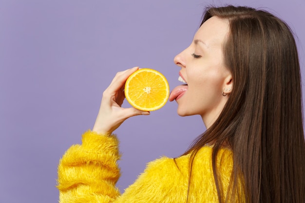 Side view of young woman in fur sweater hold licking half of fresh ripe orange fruit isolated on violet pastel background in studio. People vivid lifestyle, relax vacation concept. Mock up copy space.