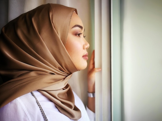 ヒジャブを着た若い女性が自宅の窓から眺めている