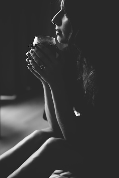 Photo side view of young woman holding drink while sitting in darkroom