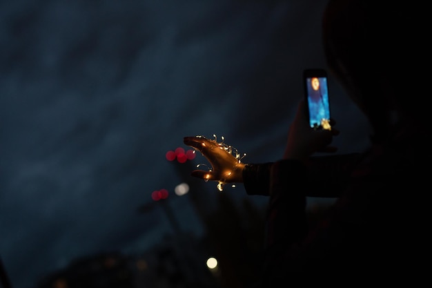 Side view of young woman holding illuminated string lights while using mobile phone against sky at night