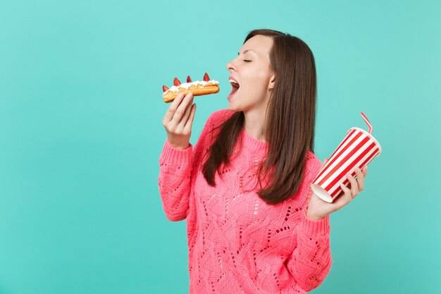 Side view of young woman in knitted pink sweater eating eclair cake hold in hand plastic cup of cola or soda isolated on blue background, studio portrait. People lifestyle concept. Mock up copy space.