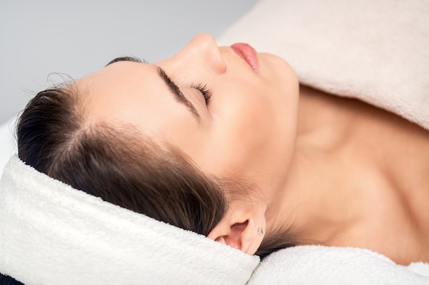 Side view of young woman lying on beautician table with closed eyes while waiting for cosmetic procedure in beauty salon