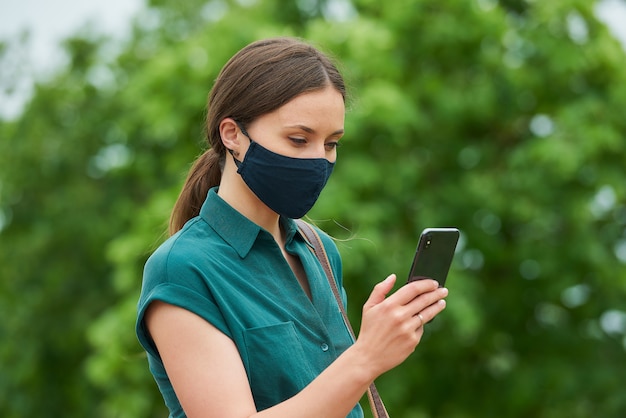 A side view of a young woman in a medical face mask is reading news on a smartphone while walking in the park