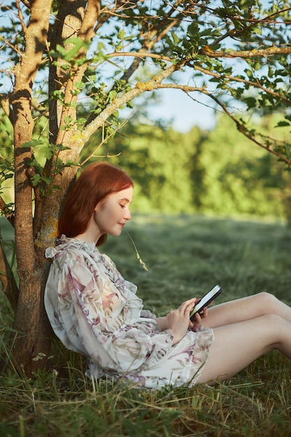여름옷을 입은 젊은 여성이 나무 아래에 앉아서 휴대폰을 보고 있는 모습
