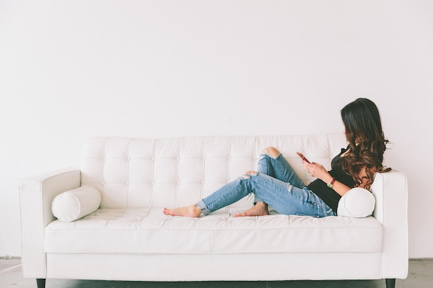 Photo side view of young woman using smart phone while sitting on sofa at home