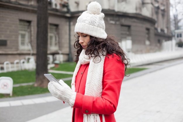 街道に立っている間スマートフォンを使って暖かい服を着た若い女性のサイドビュー