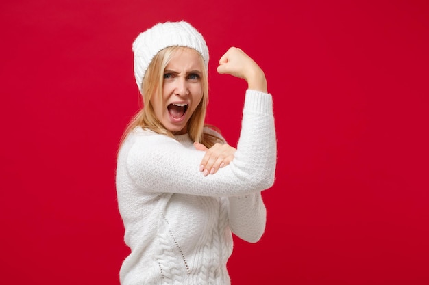 Photo side view of young woman in white sweater, hat isolated on red background studio portrait. healthy fashion lifestyle, people emotions, cold season concept. mock up copy space. showing biceps, muscles.