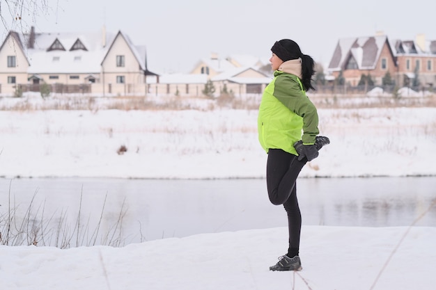 Side view of young woman with black hair stretching leg while preparing for running in winter park