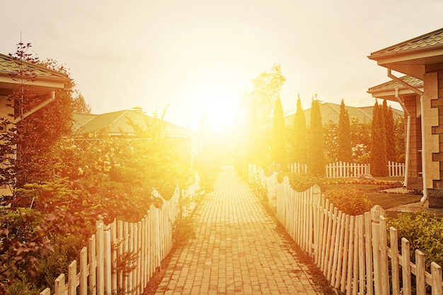 Sidewalk between cottages and gardens to rising sun small fences around road nice houses and trees s...