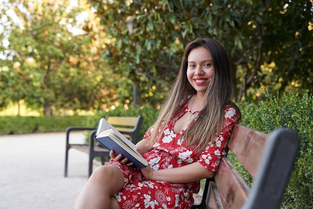 Sideways portrait of attractive schoolgirl with book looking at camera while sitting at wooden bench outdoors admiring nature