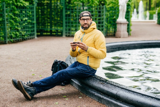 Sideways portrait of bearded male dressed in casual clothes wearing eyeglasses holding smartphone typing something looking aside having pensive expression isolated over fountain and monuments of park