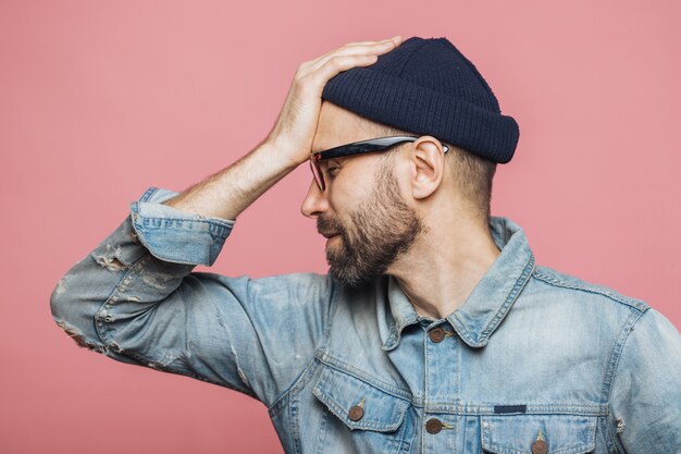 Sideways portrait of bearded male has regretful expression, keeps hand on forehead