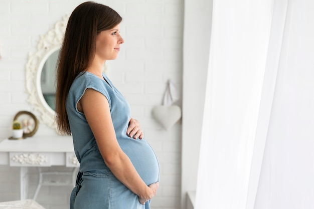 Боком беременная женщина смотрит в окно