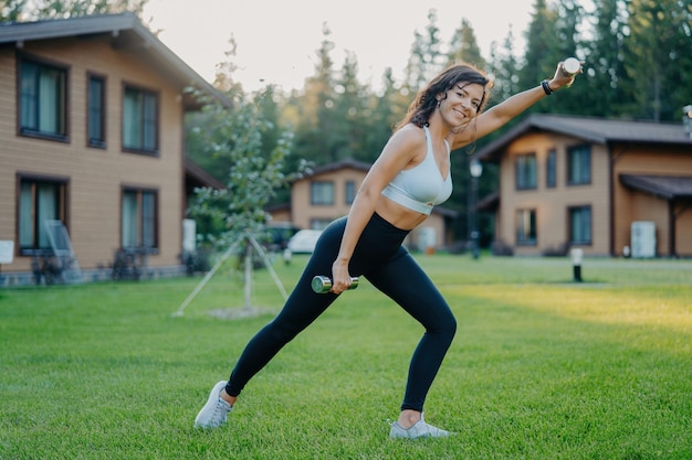 Sideways shot of active smiling determined woman raises dumbbells and has morning workout dressed in active wear poses on green lawn against private houses sportswoman trains biceps with weights