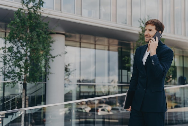 Sideways shot of bearded man calls via smartphone dressed in black formal suit poses outdoor