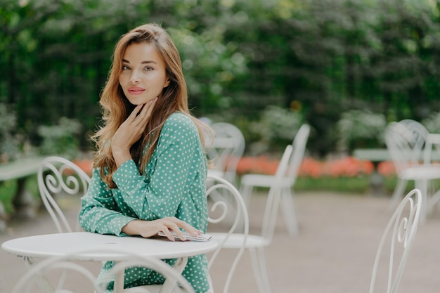 Sideways shot of beautiful caucasian woman in elegant clothes\
wears makeup holds mobile phone spends free time in outdoor cafe\
waits for friend people lifestyle rest and technology concept