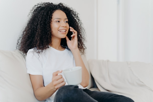 Sideways shot of positive ethnic brunette woman has telephone conversation, drinks hot beverage