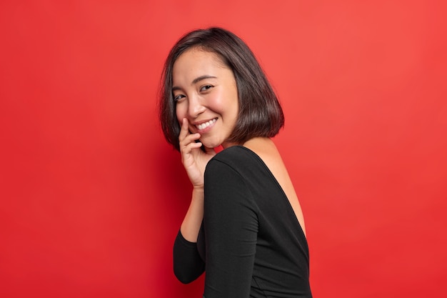 Sideways shot of pretty Asian woman smiles gently has shy happy expression wears black dress expresses positive authentic emotions isolated over vivid red wall