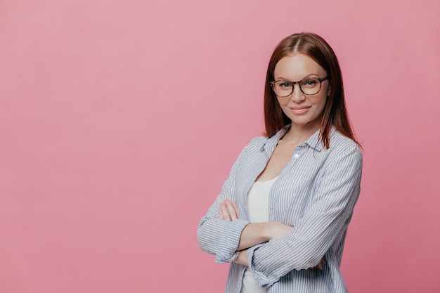 Sideways shot of prosperous businesswoman keeps arms folded, dressed in formalwear, wears spectacles