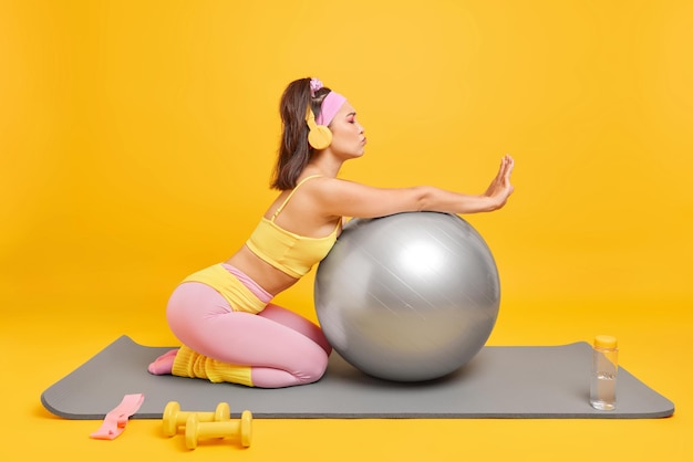 Sideways shot of slim brunette young woman leans on fitness ball poses on karemat listens music via headphones during sport training isolated over yellow background. Wellness and pilates concept