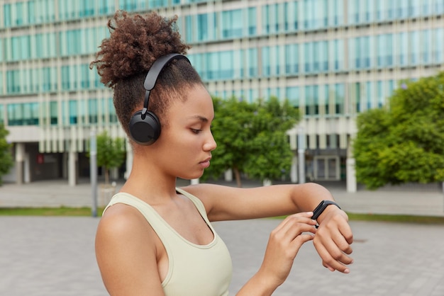 Sideways shot of sporty woman dressed in t shirt checks fitness results on smartwatch listens motivational music via headphones poses against urban background athletic female model goes in for sport