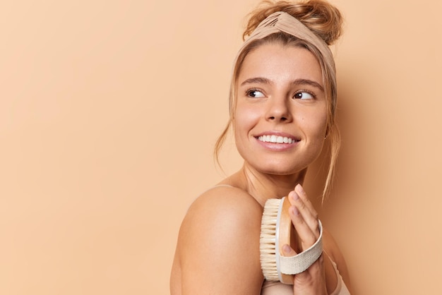 Sideways shot of tender cheerful woman uses body brush stands half naked in profile wears headband isolated over beige background with copy spce for your promotional content or advertisement