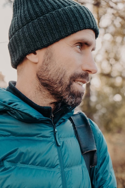 Sideways shot of unshaven man with dark thick beard wears warm\
har and anorak looks pensively aside poses outside dreams about\
something has outdoor stroll in unknown place alone active\
lifestyle