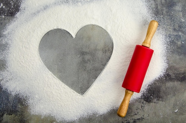 Sifted scattered flour and rolling pin on a dark background of a concrete table top. Floured cutting board. Space for text in the shape of a heart. Grainy surface, selective focus.