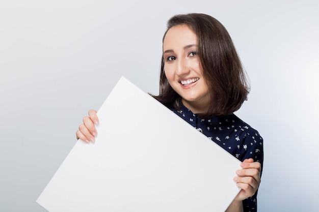 Sign board. Business woman holding big white card. Isolated portrait. Young happy, smiling girl holding a blank sheet of paper for advertising