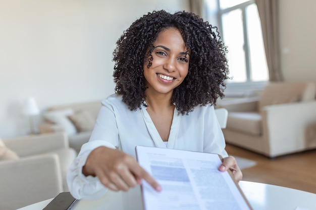 Firmi qui giovane donna sorridente fiduciosa che punta a un foglio di carta contratto in possesso di una penna guardando e mostrandolo alla fotocamera concetto di business accordo di presentazione