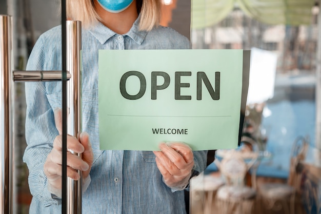 Photo sign open welcome on shop entrance door as new normal. young woman in protective medical mask and gloves hangs on front door of cafe reopen sign. end lockdown coronavirus covid 19 for local business.