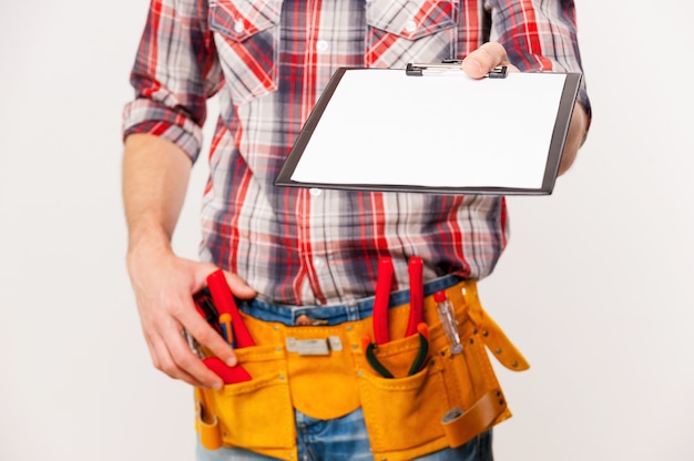 Sign this. Close-up of handyman with tool belt stretching out clipboard with paper standing against grey background