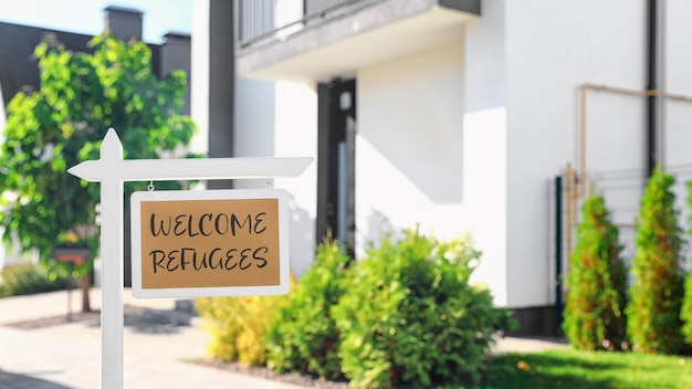 Sign with phrase WELCOME REFUGEES near house outdoors on sunny day