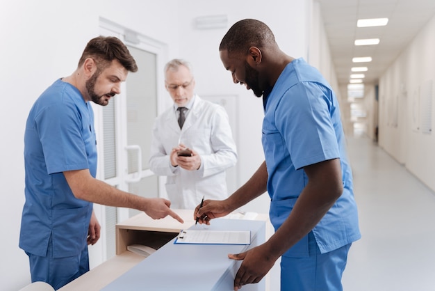 Signing important document . Cheerful skilled diligent doctors working in the hospital and discussing diagnosis of the patient while aged colleague using phone in the background