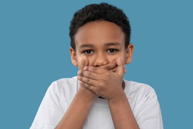 Silence. Cheerful dark-skinned boy in white tshirt covering his mouth with palms standing in studio photo