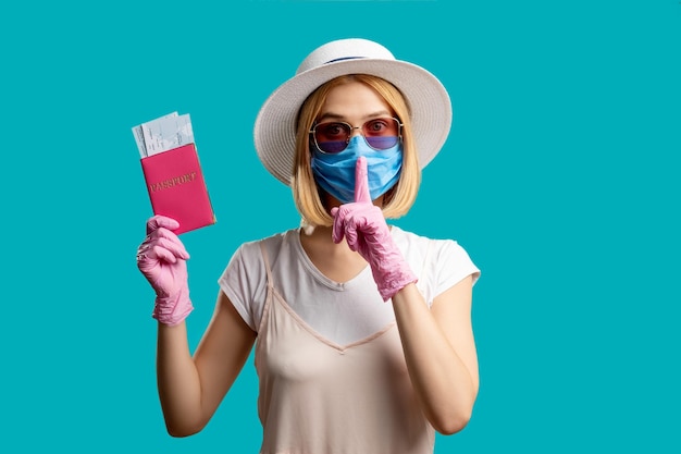 沈黙のジェスチャー旅行制限保護マスクの女性ピンクの手袋をはめたパスポートを持ってチケットを口に指を突き刺す青で隔離されたカメラを見て秘密を守る検疫防止