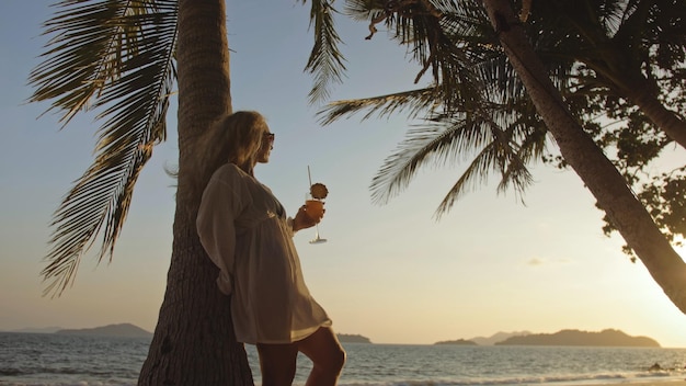 SILHOUET DONKERGOUD ZONSONDERGANGZICHT Mooie vrouw die ananascocktail Pina Colada drinkt dichtbij palmboom Vrouw in wit overhemdszonnebril Concept rust tropische zee reizend toerisme zomervakantie