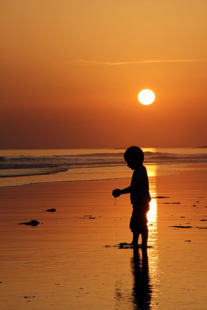 Silhouet jongen staat op het strand tegen de oranje hemel tijdens de zonsondergang