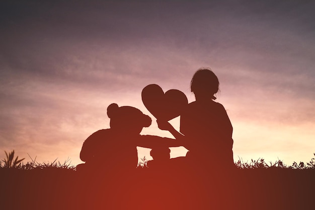 Silhouet meisje met teddybeer en hartvorm op het veld tegen de lucht