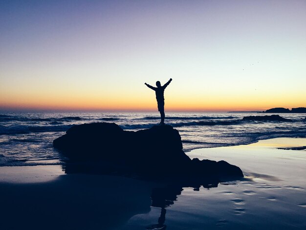 Foto silhouet persoon met uitgestrekte armen die tijdens de zonsondergang op een rots op het strand staat