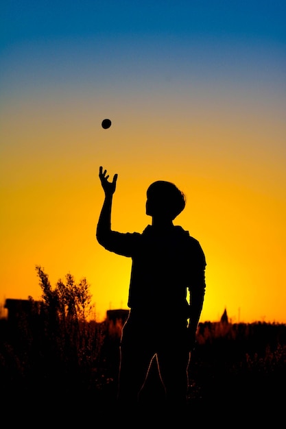 Foto silhouet tienerjongen die met een bal speelt terwijl hij tegen de hemel staat tijdens de zonsondergang