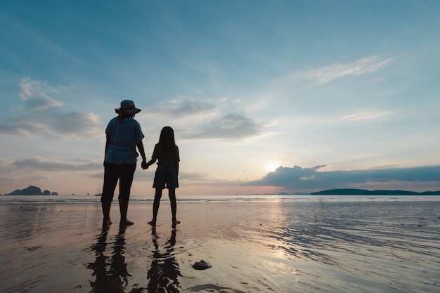 Silhouet van Aziatische moeder en dochter die hand samen houden die zich op het strand bevinden die de mooie overzees en de hemel bekijken in de zonsondergangtijd.