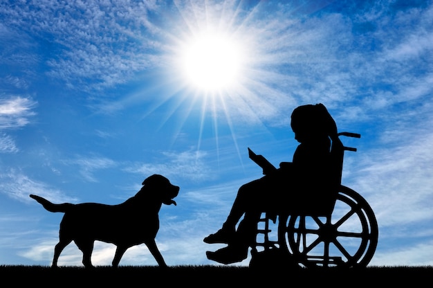 Silhouet van een gehandicapt kindmeisje dat in een rolstoel zit en een boek leest in de buurt van haar hond. Conceptueel beeld van het leven van kinderen met een handicap