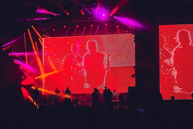 Silhouet van een grote menigte tijdens een concert tegen een helder verlicht podium. Nachtelijk rockconcert met mensen die plezier hebben en hun handen in de lucht steken en de muzikanten toejuichen