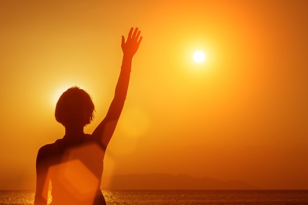 Silhouet van een jong meisje dat op het strand staat met de hand omhoog bij de zonsondergang tegen de zon