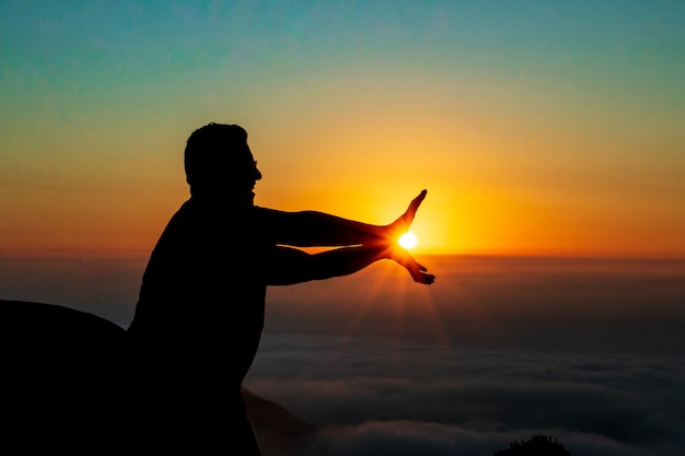silhouet van een man die de zon aanraakt met beide handen op de top van een berg