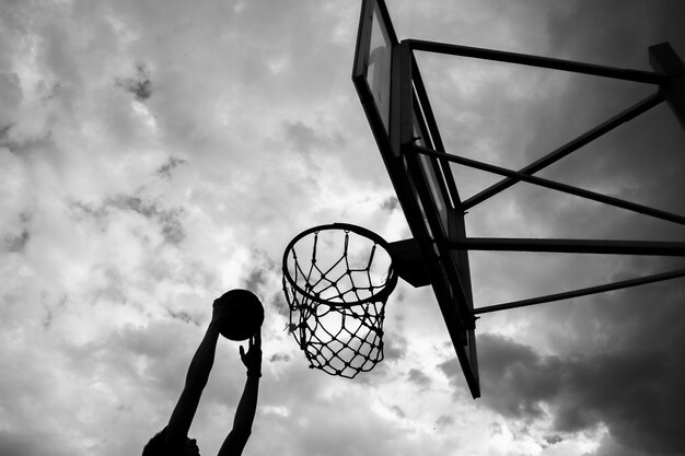 Silhouet van een man die een bal in een basketbalring gooit op straat tegen een lucht met wolken in zwart-witte kleur